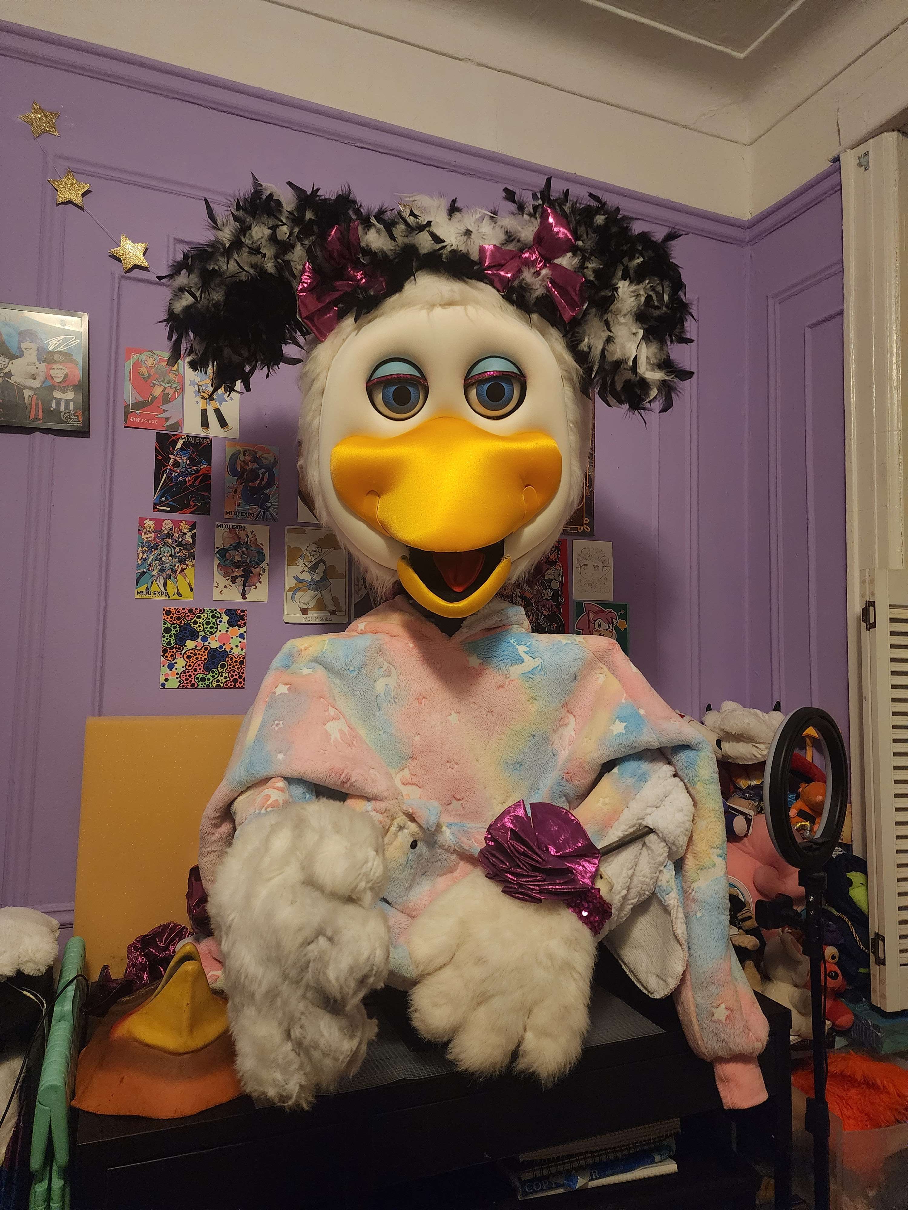 my helen animatronic. she's sitting on a black desk in front of a purple wall covered with art. she has a pastel pink and blue snuggie draped over her shoulders.